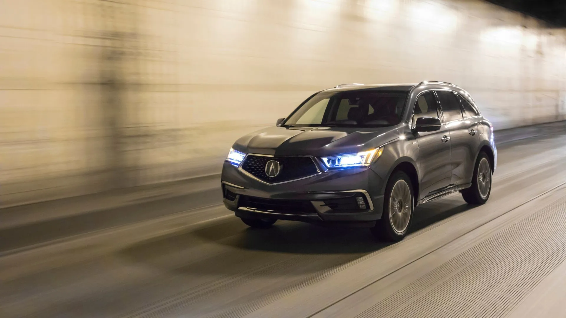 Hình ảnh tổng thể xe SUV Acura MDX Sport Hybrid 2020 màu xám đang di chuyển trong hầm với đèn pha bật sáng, nhấn mạnh tầm quan trọng của hệ thống chiếu sáng xe.