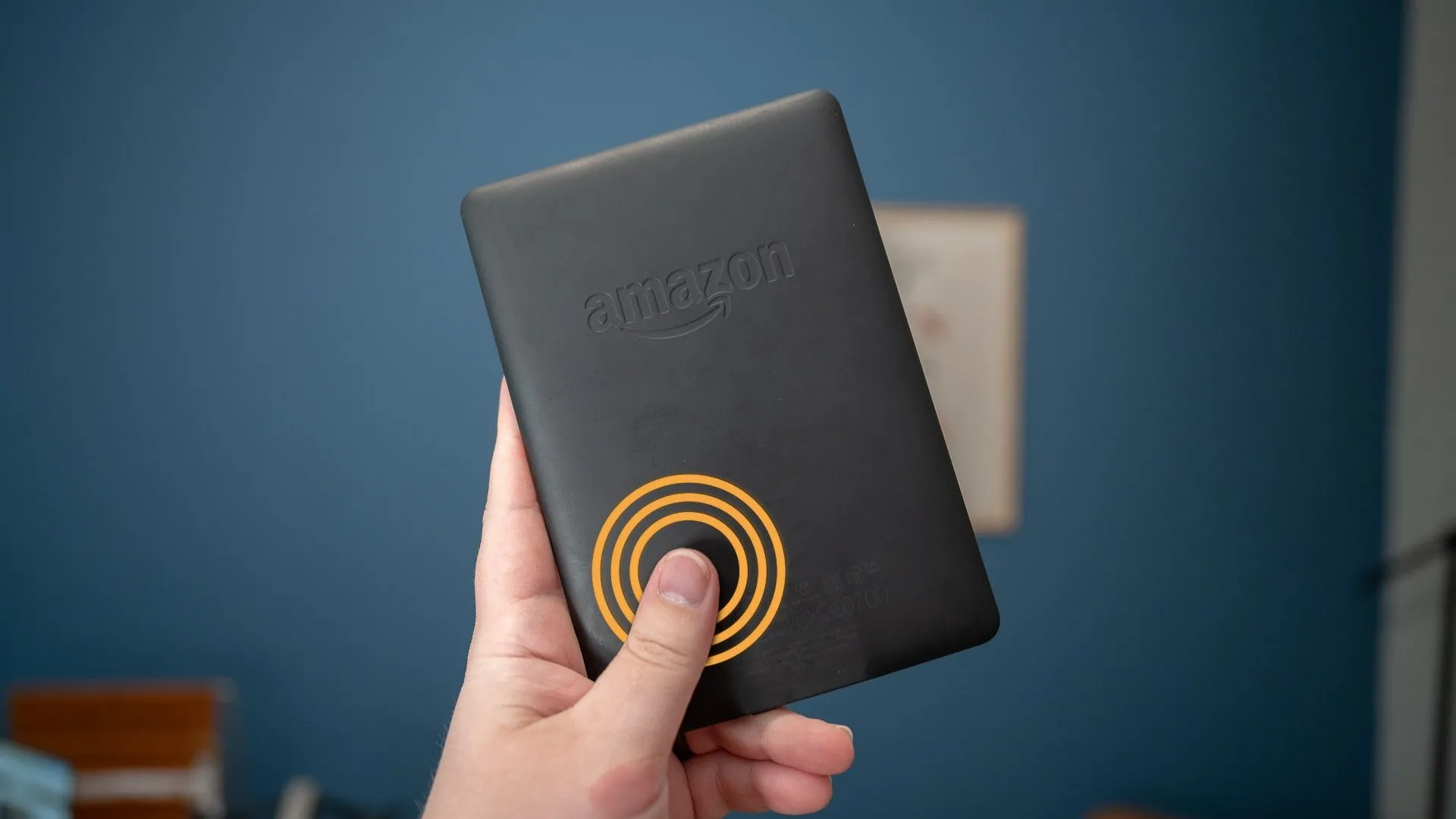 Người dùng đang cầm thiết bị đọc sách Kindle Paperwhite hoặc Kindle Colorsoft, với ngón tay chạm vào mặt lưng để thực hiện cử chỉ lật trang
