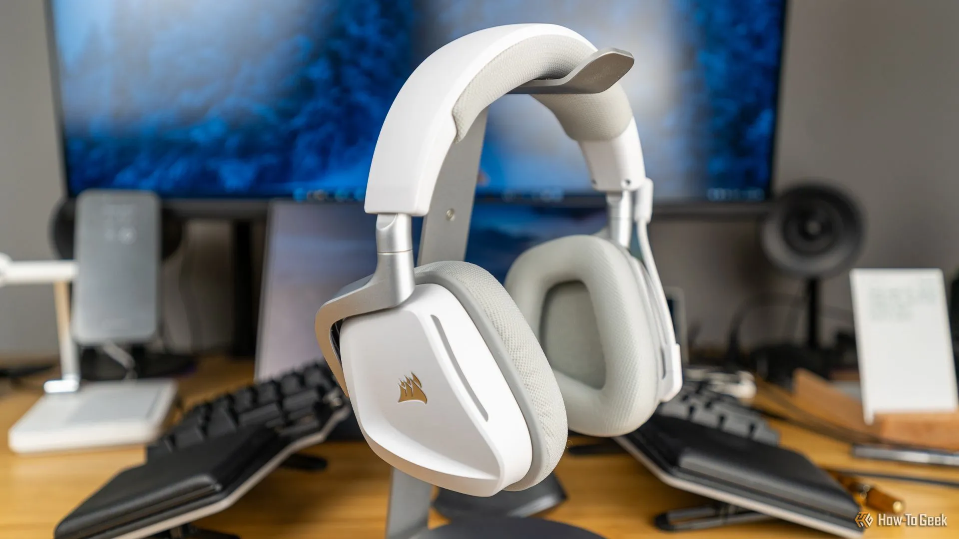 Tai nghe gaming Corsair Void Wireless V2 màu trắng đặt trên giá đỡ tai nghe, cùng các phụ kiện gaming khác trên bàn làm việc.